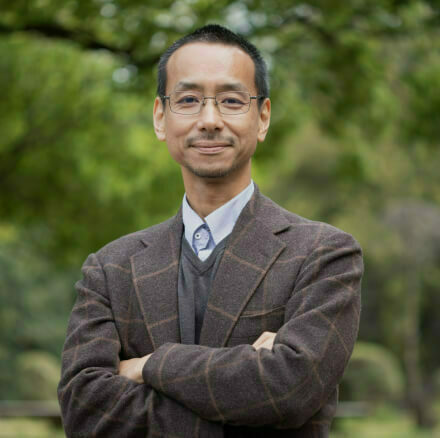 Headshot of Junichiro Shiomi. An East Asian man with short dark hair and a light mustache and goatee, smiling slightly. He wears a tan suit jacket, a white collared shirt, and a striped tie against a gray background.